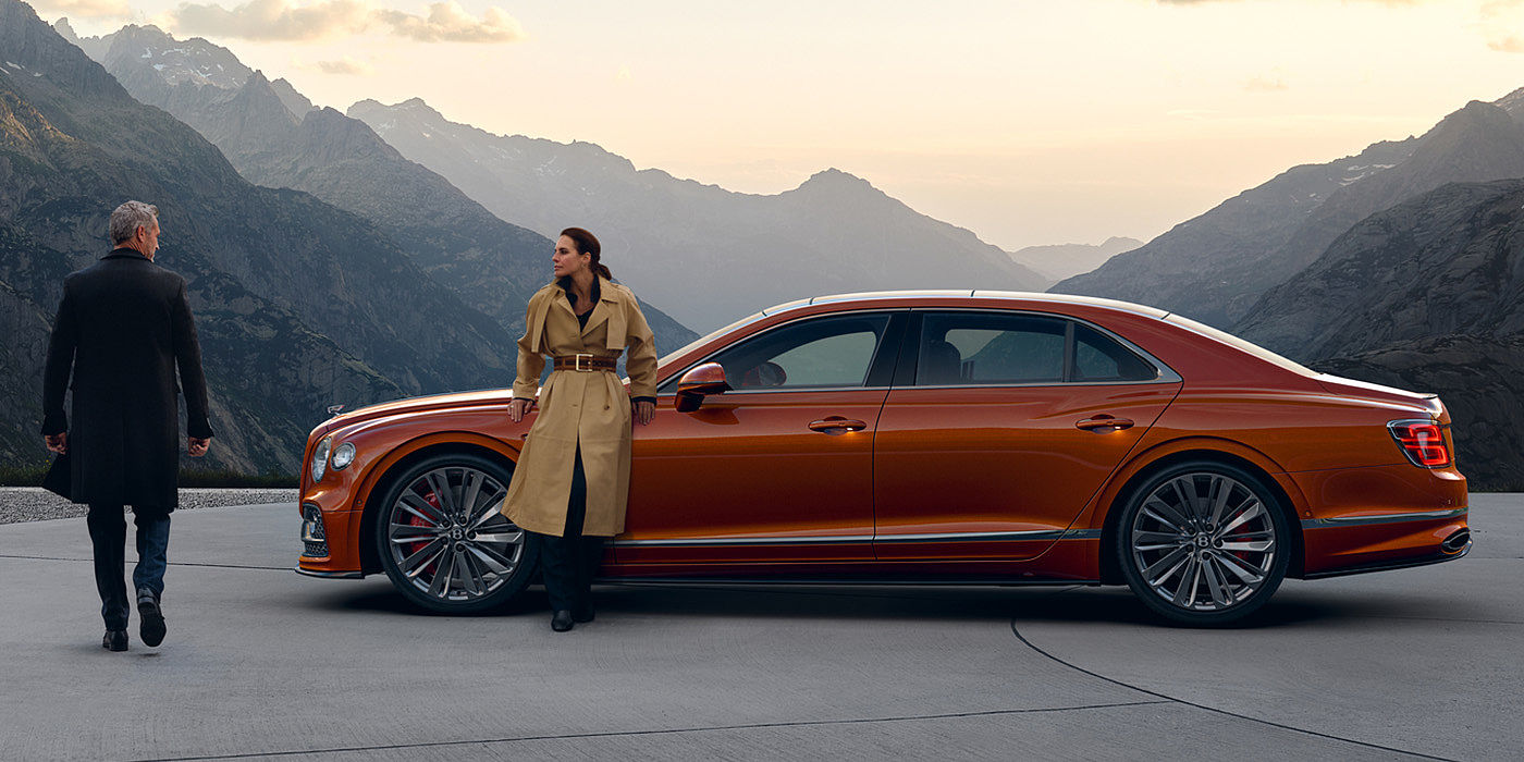 Bentley Valencia Bentley Flying Spur Speed parked in Orange Flame coloured exterior parked, with mountainous background and two people in view.