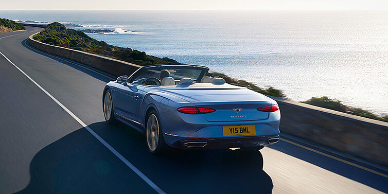 Bentley Valencia New Bentley Continental GTC Azure convertible rear view in Light Windsor blue paint with 22 inch ten spoke wheels driving dynamically on an ocean road in the sunshine