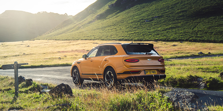 Bentley Valencia Bentley Bentayga S SUV rear three quarter in Sunburst Gold paint on a road surrounded by green hills