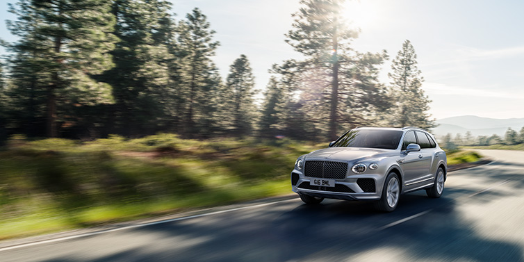 Bentley Valencia Bentley Bentayga SUV front three quarter in Moonbeam silver paint driving dynamically along a tree-lined road