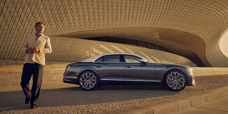 Bentley Valencia Bentley Flying Spur Mulliner sedan in Rose Gold over Magnetic duotone paint parked by a curved wall with person in suit jacket standing on the left